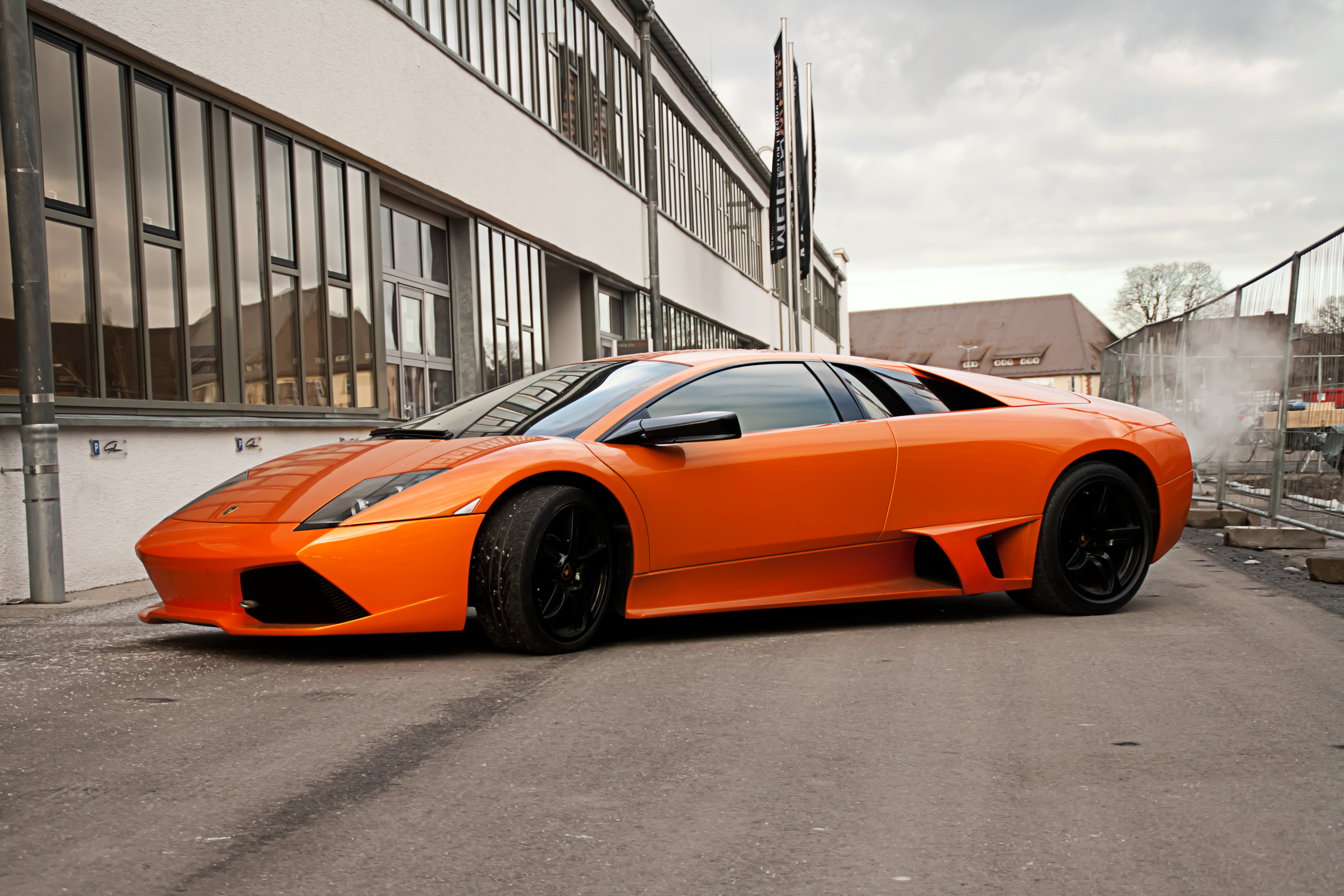 Скачать lamborghini, murcielago, orange, lp640, ламборгини, мурсиэлаго, оранжевый, вид сбоку, чёрные диски, здание, окна, ограждение, небо, тучи обои на рабочий стол